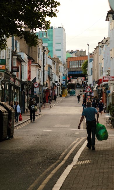 A view of a city street in Dulwich showing a variety of commercial and residential buildings with mixed architectural styles, including brick facades and modern glass structures in the background. The street has a gentle incline and is lined with shops, some of which have signage and awnings. Pedestrians are walking along the pavement, with some carrying shopping bags, and a man in the foreground walks away from the camera holding a large green waste bag. The scene is captured in natural daylight, with overcast skies providing diffuse lighting. Trees with green foliage partially frame the upper part of the image. The street appears to be a mix of pedestrian and vehicle use, with white road markings visible on the tarmac, and a few small parked vehicles can be seen further along the street. This setting exemplifies a typical urban environment where private waste collection services, such as those provided by Waste Collection Dulwich, might be involved with rubbish removal or on-site clearance, especially in the context of supporting frequent or alternative waste handling methods for local residents and businesses.