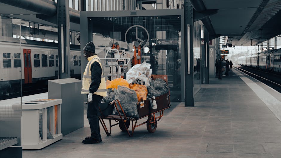 A man wearing a dark beanie, glasses, and a high-visibility vest stands on a concrete platform next to a red, metal wagon filled with various piles of waste materials wrapped in plastic and fabric bags, some in orange and black colors. The wheeled cart is positioned near an outdoor train station with a glass shelter behind him, which houses maintenance equipment and buffers. To the left, a white waste bin with a lid is situated against the station's metallic structure. The background features train tracks with a moving train on the right side, and distant station platform figures are visible. The environment is well-lit by natural daylight, highlighting the textures of the waste materials, the smooth surfaces of the station, and the reflection of the surroundings in the glass shelter. The scene appears to relate to private waste collection or on-site clearance services, showing an individual managing rubbish at a public transport hub, possibly associated with waste management services like Waste Collection Dulwich or broader rubbish removal procedures in urban environments.