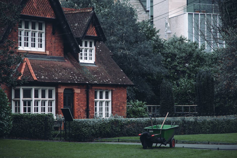 The image displays a urban residential scene with a traditional red-brick house featuring a steeply pitched roof, dark tiles, and white-framed sash windows. The house is situated behind a low, neatly trimmed hedge and has a small, enclosed front garden area. In the foreground, a green wheelbarrow with red wheels is positioned on a paved pathway, with two shovels resting inside it. To the right of the wheelbarrow, a black waste bag is placed on the ground. The surrounding environment includes well-maintained grassy lawns and trees, with some dense foliage on the right side, partially obscuring a modern taller building in the background. The overall lighting suggests an overcast day, contributing to a subdued and neutral atmosphere that aligns with the context of managing and disposing of garden or household waste, often managed by private waste collection services such as waste collection Dulwich.