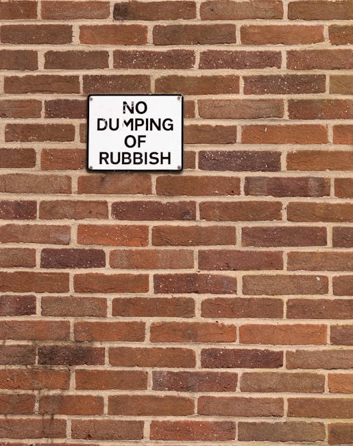 A rectangular white metal sign with black uppercase lettering reading 'NO DUMPING OF RUBBISH' is mounted on a red-brown brick wall with a traditional running bond pattern. The wall features a mix of warm reddish tones and darker brown bricks, with light grey mortar joints creating a uniform horizontal and vertical grid. The sign is positioned slightly above the center of the image, with its edges aligned neatly with the brickwork. The lighting appears natural and even, casting minimal shadows and highlighting the texture of the brick surface. The scene suggests a maintained exterior environment, potentially near a private property or commercial premises, where waste management and rubbish disposal are regulated, aligning with independent local waste collection practices overseen by waste management services like Waste Collection Dulwich, which handle rubbish removal and clearance for residential and commercial clients.