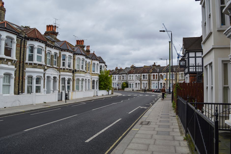 A residential street scene featuring a row of Victorian-style terraced houses with white and brick facades, decorative bay windows, and clay tile roofs under an overcast sky. The houses are situated along a paved sidewalk with a black metal fence and small garden spaces in front. To the right, a white building with large windows and traditional architecture is visible. The street has clearly marked parking bays and a single lane for vehicles, with a few cars parked along the curb. In the background, a construction crane indicates ongoing development or renovation work on nearby buildings. The overall atmosphere is quiet and urban, with natural light diffused by the cloudy sky, typical of a Dulwich neighborhood. This scene subtly relates to private waste management, as properties in this area may require alternative rubbish collection and disposal services provided by companies like Waste Collection Dulwich, especially for out-of-hours or on-site clearance needs.
