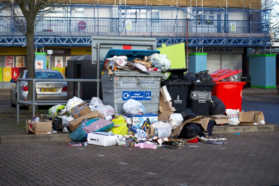 An outdoor scene showing a large pile of mixed rubbish and waste materials overflowing from multiple garbage bins in a public parking area. The waste includes cardboard boxes, plastic bags, paper packaging, and other household refuse, some of which has spilled onto the brick-paved ground surrounding the bins. There are at least four visible bins; one of grey color designated for mixed paper and cardboard, a black bin likely for general waste, a red bin for recyclables, and another black bin partially obscured. The waste appears uncontained, with loose items and bags scattered across the immediate area. In the background, there is a modern commercial building with a blue scaffolding structure and storefronts, including a fish bar. A parked silver car is behind a metal railing on the left, partially blocking the view of the building. The scene suggests an instance of overflowing rubbish at an urban location, illustrating the need for regular waste collection and disposal services, such as those provided by Waste Collection Dulwich for private or independent waste handling in the area.