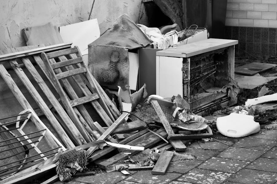 A collection of discarded household items and waste materials spilling onto a paved outdoor area, including a leaning wooden garden chair with slatted backrest, weathered and aged, situated on the left side of the image. Behind it, a partially dismantled piece of furniture or appliance features a damaged, exposed interior, with chipped paint and worn surfaces. Nearby, there are broken wooden planks, scattered debris, and a damaged white plastic object resembling a toilet seat or lid lying on the ground. In the background, a brick wall and a section of concrete or stone surface create an urban setting. The environment appears dirty and neglected, with various textures and materials such as wood, plastic, and metal visibly mixed, characteristic of a collection prepared for disposal or private rubbish removal. This scene exemplifies typical clutter that waste removal services, like those offered by Waste Collection Dulwich, may handle as part of on-site clearance or private waste collection efforts in East Dulwich, Dulwich.