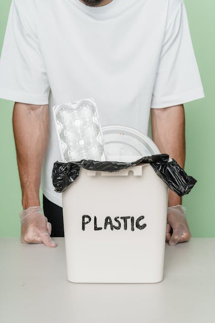 A black wheeled rubbish bin with a white label and the name 'St. John’s' printed on the front, positioned on a paved sidewalk adjacent to a curb in a residential or urban area at night. The lid is open, revealing a mix of waste materials including cardboard boxes, paper bags, and plastic packaging, which are spilling out slightly. In the background, there are streetlights illuminating a quiet street lined with trees and houses, with some vehicle lights visible at a distance. The environment suggests an informal waste collection setting, potentially representing an alternative or private rubbish disposal method, as the bin appears to be designated for a specific property or community. The scene conveys a typical scene of rubbish awaiting collection, aligned with waste management services, with natural evening lighting emphasizing the everyday urban waste collection process.